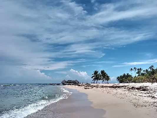 Strandblick von Playa Hemingway in Juan Dolio mit klarem Himmel, sanften Wellen, Palmen und einem traditionellen Gebäude am Horizont.